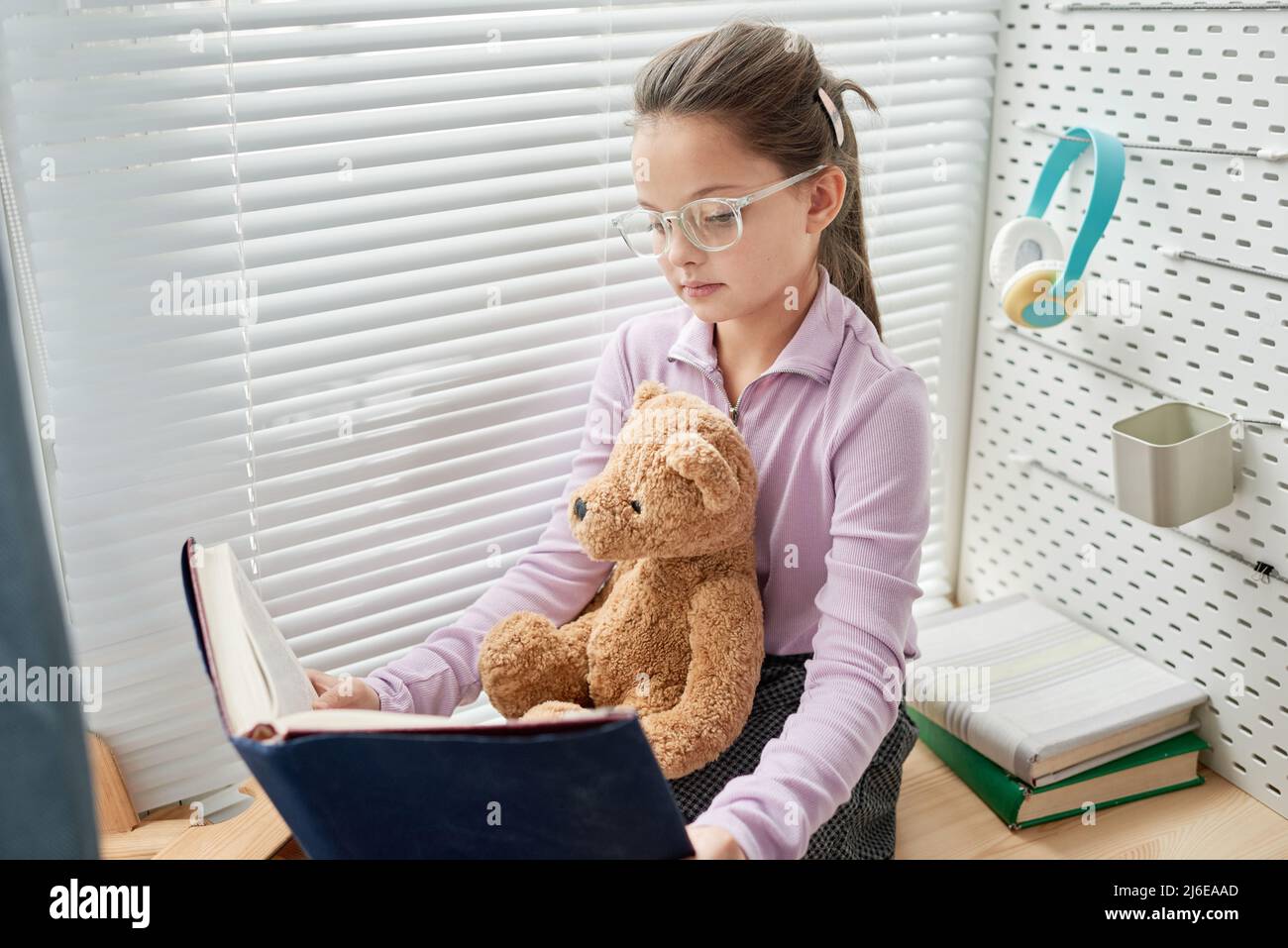 Konzentrierte kluge Mädchen in Gläsern sitzen mit Spielzeug Bär auf den Knien und Buch zu lesen, um es Stockfoto