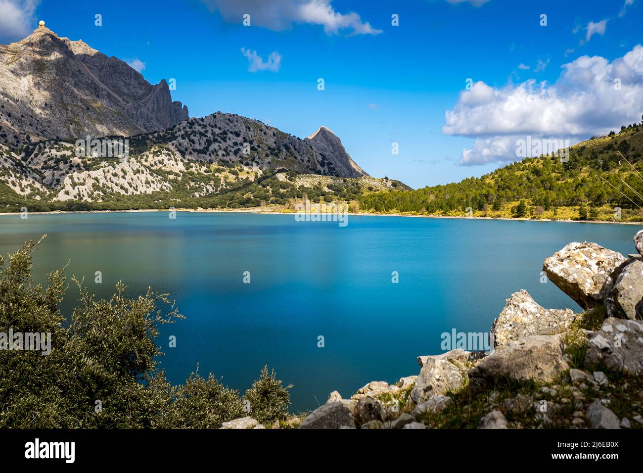 Künstlicher Bergsee auf Mallorca mit Felsen und Busch im Vordergrund und den Bergen Puig Major und Es pa de Figa im Hintergrund. Stockfoto