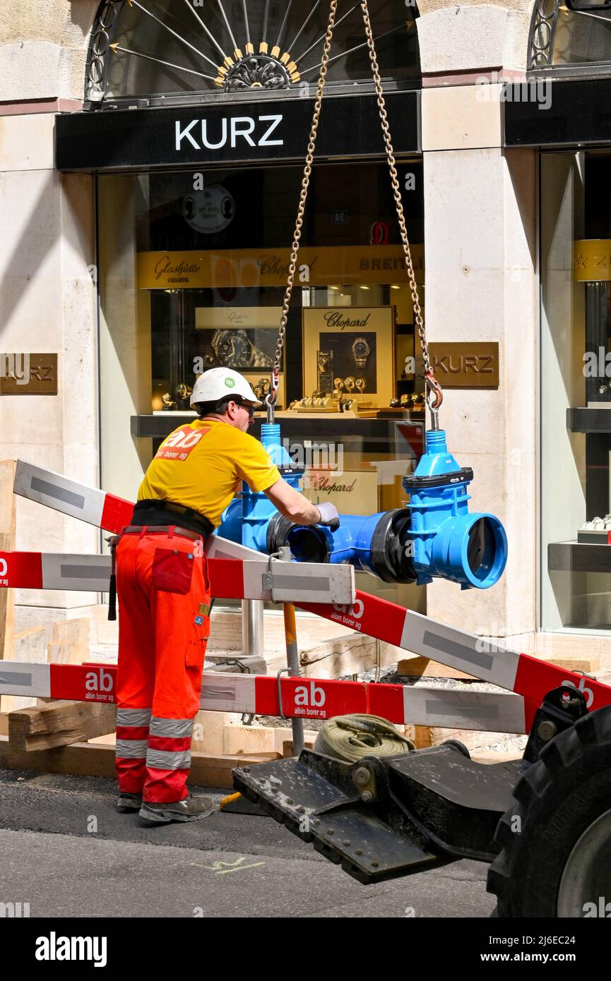 Basel, Schweiz - April 2022: Bauarbeiter hilft, eine schwere Rohrverbindung in einen Graben in einer Straße im Stadtzentrum zu senken Stockfoto