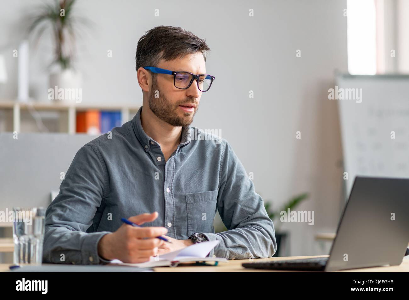 Erfolgreicher Geschäftsmann mittleren Alters, der im Büro arbeitet, am Schreibtisch vor dem Laptop sitzt und sich Notizen macht Stockfoto