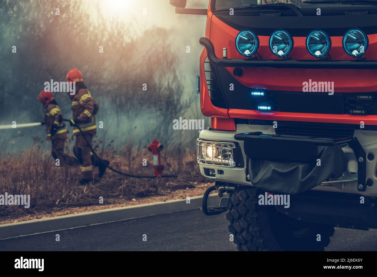 Moderner Europäischer Feuerwehrwagen und zwei Feuerwehrmänner kämpfen Grass Wildfire im Hintergrund Stockfoto