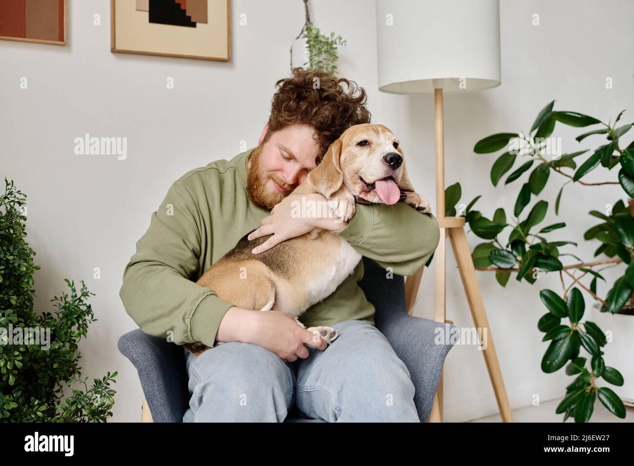 Junger bärtiger Mann, der auf einem Sessel im Zimmer sitzt, mit Pflanzen, die seinen Hund umarmen und sich entspannen Stockfoto