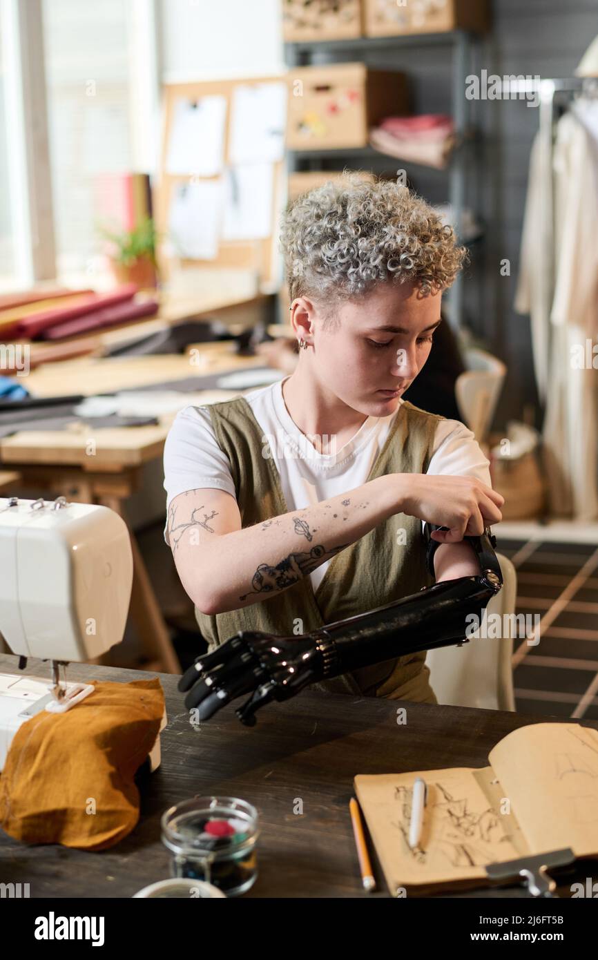 Moderne junge Frau, die während der Arbeit eine myoelektrische Armprothese anpasst, während sie am Tisch vor der elektrischen Nähmaschine sitzt Stockfoto