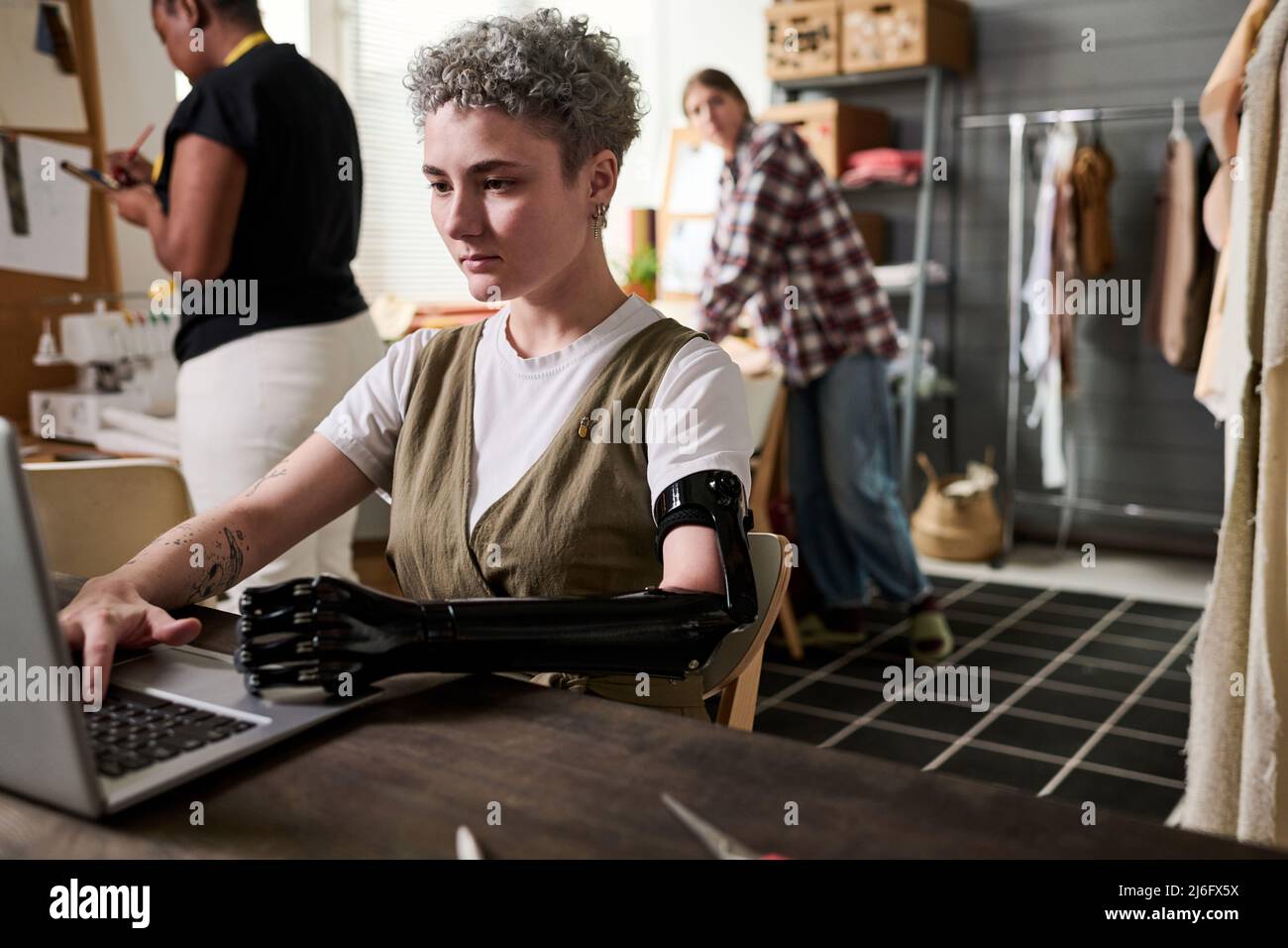 Junge, seriöse Modedesignerin mit myoelektrischem Arm, die auf dem Laptop-Tastenfeld tippt, während sie nach Online-Informationen über neue Trends sucht Stockfoto
