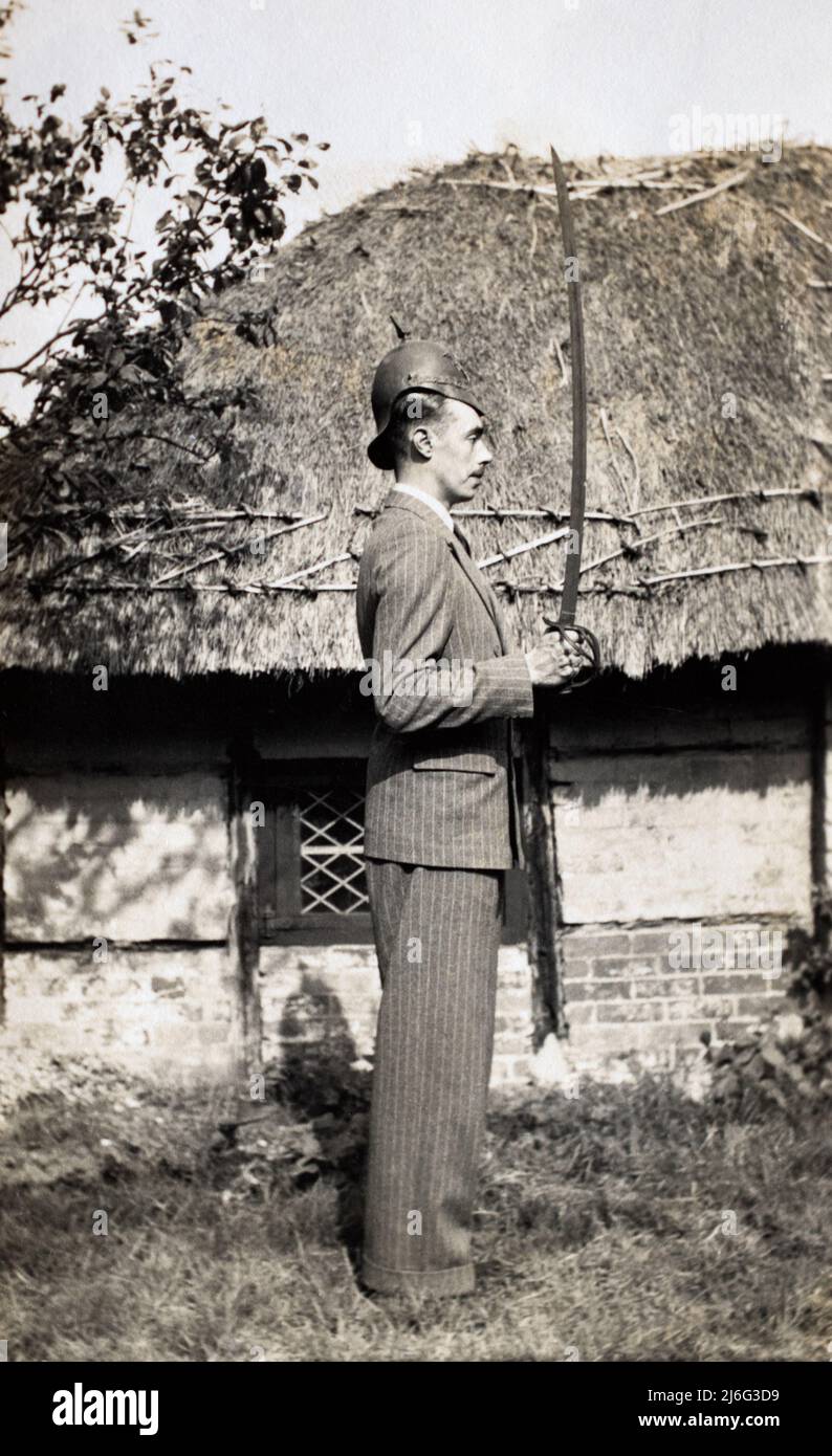 Ein Mann in einem Nadelstreifenanzug, der vor einer strohgedeckten Hütte posiert. Er trägt einen alten Metallhelm eines britischen schweren Kavallerieregiments und hält ein Schwert in der Hand. Stockfoto