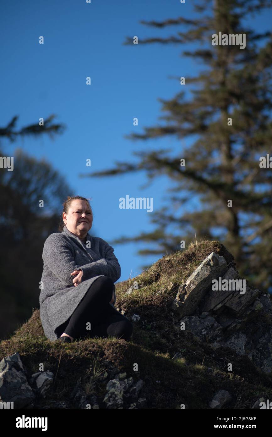Frau in grauer Strickjacke, die auf moosbedeckten Felsen unter blauem Himmel in Schottland sitzt Stockfoto