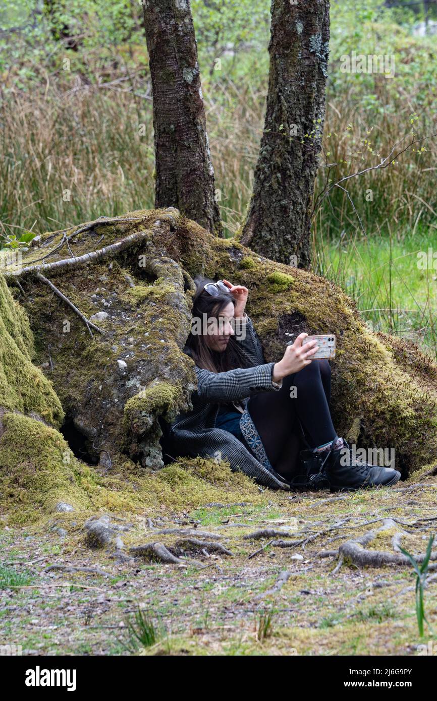 Junge Frau, die Selfie macht, während sie auf moosbedeckten Steinmauern in Schottland sitzt Stockfoto