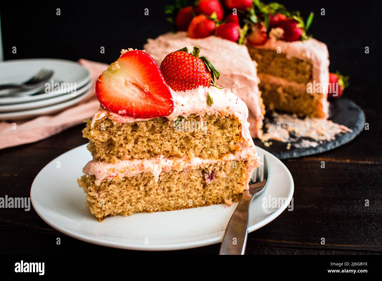 Scheibe frischer Erdbeerkuchen auf einem weißen Teller: Eine Scheibe Kuchen auf einem kleinen Teller mit dem Rest des Kuchens im Hintergrund Stockfoto