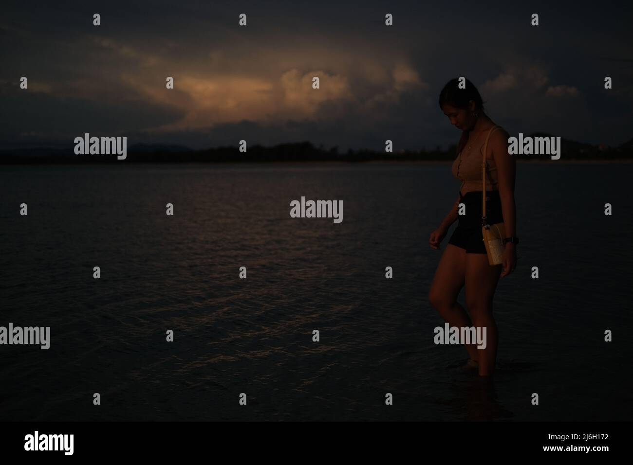 Eine junge Frau läuft knöcheltief im Wasser, vor einer szenischen Gewitterwolke, die von der untergehenden Sonne beleuchtet wird Stockfoto