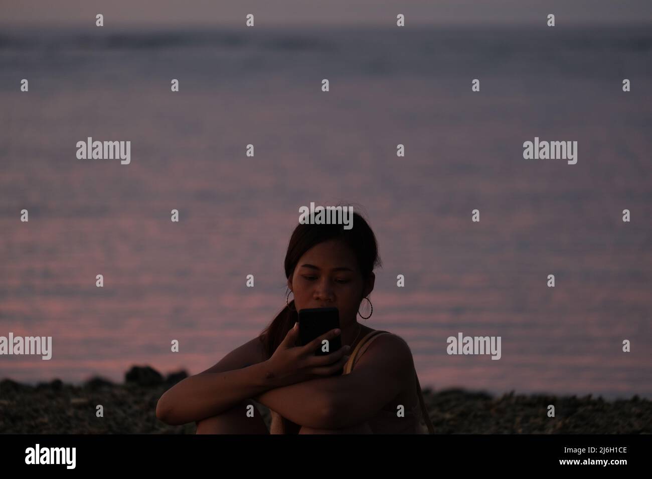 Die junge Frau nutzt das Smartphone, während sie am späten Abend an einem felsigen Strand vor dem ruhigen Meer sitzt Stockfoto