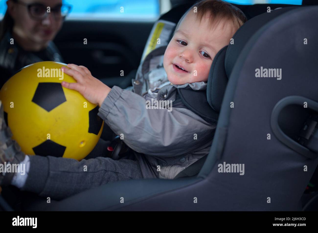 Netter kleiner Junge, der in einem Auto in seinem Stuhl sitzt und einen Ball hält Stockfoto