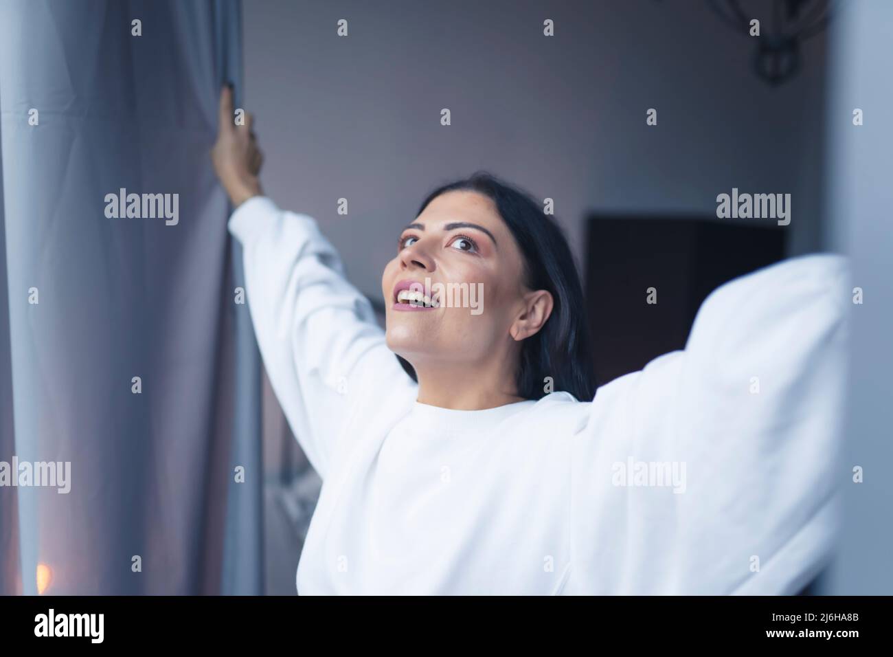 Fröhliche kaukasische schwarzhaarige Frau mit 30s in einem weißen Sweatshirt, die Fenstervorhänge öffnet und den neuen Tag begrüßt. Hochwertige Fotos Stockfoto