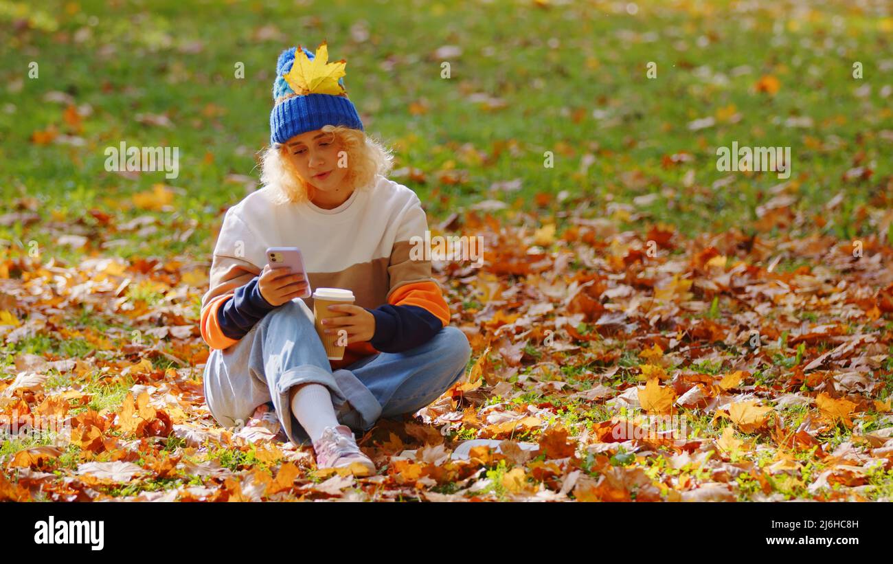 Freundliches kaukasisches blondes Mädchen, das auf einem Boden sitzt, der mit bunten, trockenen Herbstblättern bedeckt ist, eine Tasse Kaffee zum Mitnehmen hält und durch ihr Telefon scrollt. Konzept für Freizeitaktivitäten. Hochwertige Fotos Stockfoto