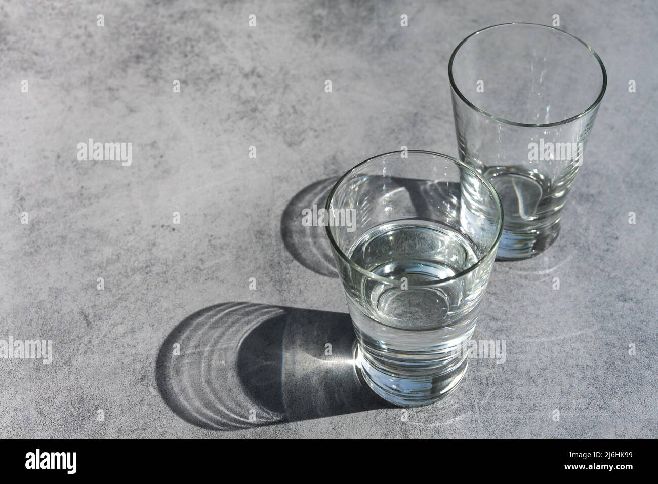Trinkgläser, die eine Hälfte mit Wasser gefüllt, die andere leer, mit Schatten und Lichtreflexen auf einem rustikalen grauen Hintergrund, Konzept für Klima Cha Stockfoto