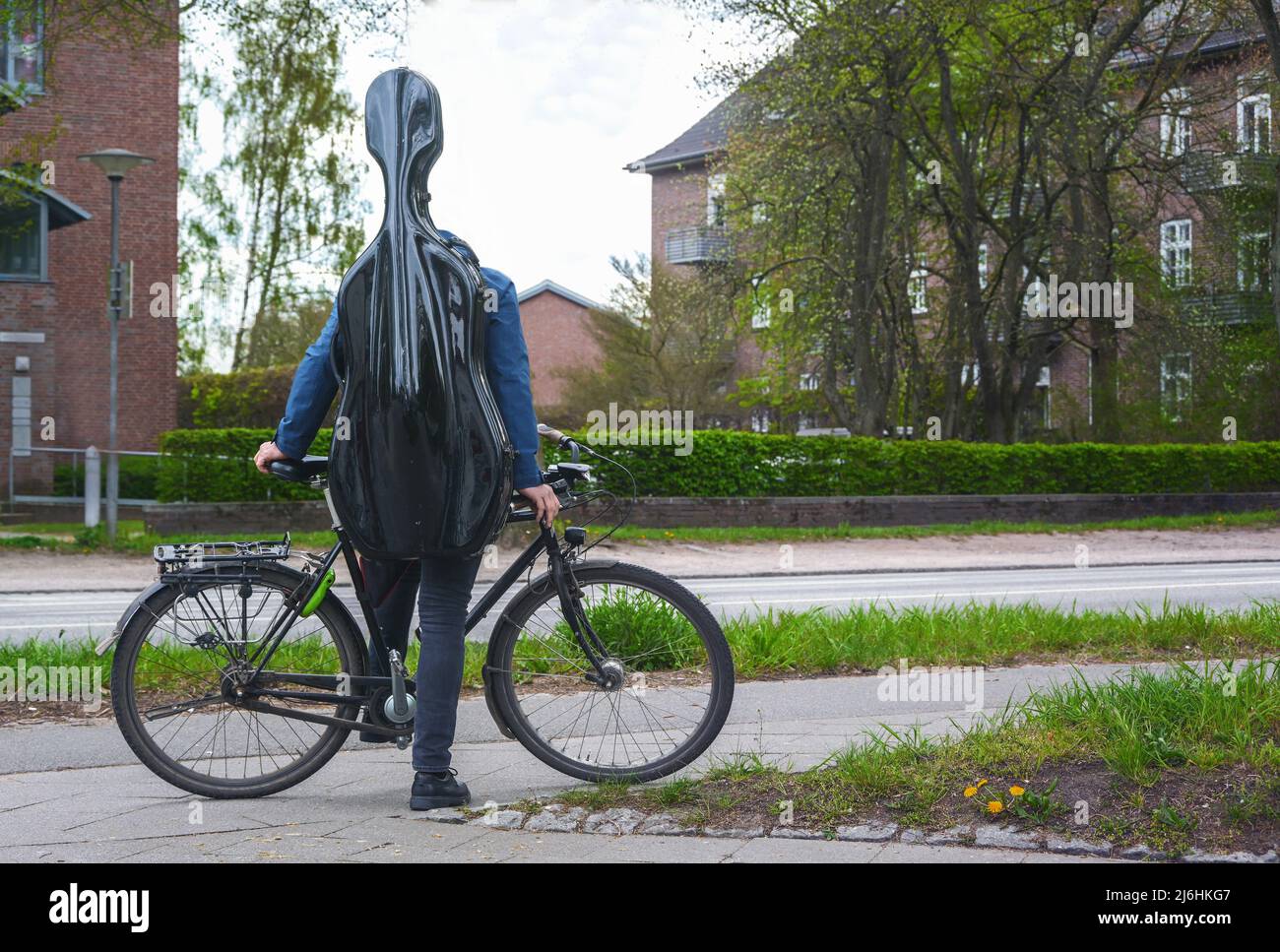 Der junge Musiker mit einem schwarzen Cellogehäuse auf dem Rücken steht mit seinem Fahrrad in einer Vorstadtstraße in einem Wohngebiet auf dem Weg zum Musikunterricht oder t Stockfoto