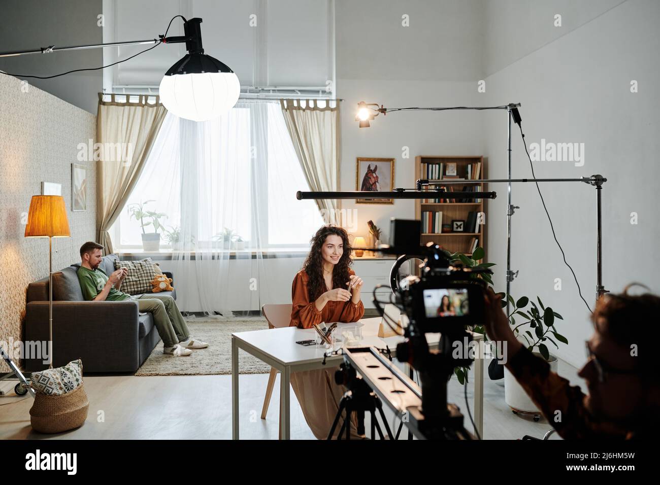 Glückliche junge Bloggerin, die am Tisch in einer Loft-Wohnung sitzt, während der Videoaufzeichnung, dass der Werbespot vom Kameramann aufgenommen wurde Stockfoto