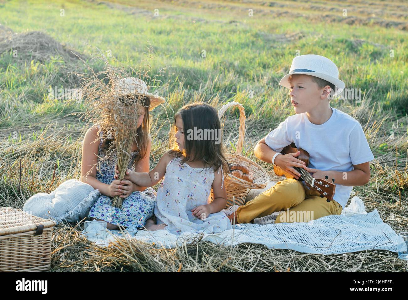Porträt eines fröhlichen Jungen und zwei Mädchen, die auf einer Decke auf dem Feld sitzen und ein Picknick machen. Erholsame Zeit. Gitarre spielen, Ukulele spielen, reden, scherzen. Leicht sonnig Stockfoto