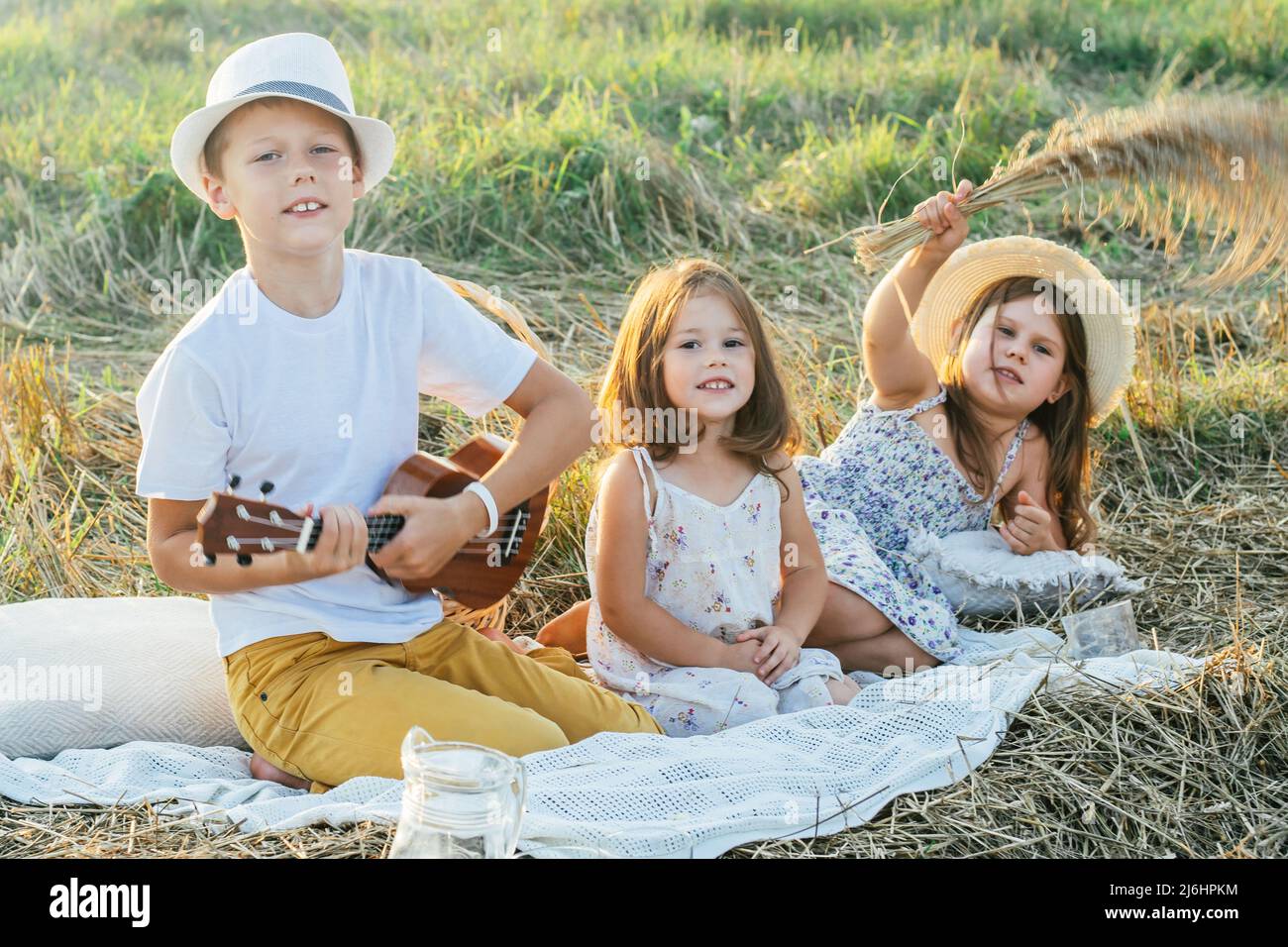 Porträt eines fröhlichen Jungen und zwei Mädchen, die auf einer Decke auf dem Feld sitzen und ein Picknick machen. Erholsame Zeit. Gitarre spielen, Ukulele spielen, reden, scherzen. Leicht sonnig Stockfoto