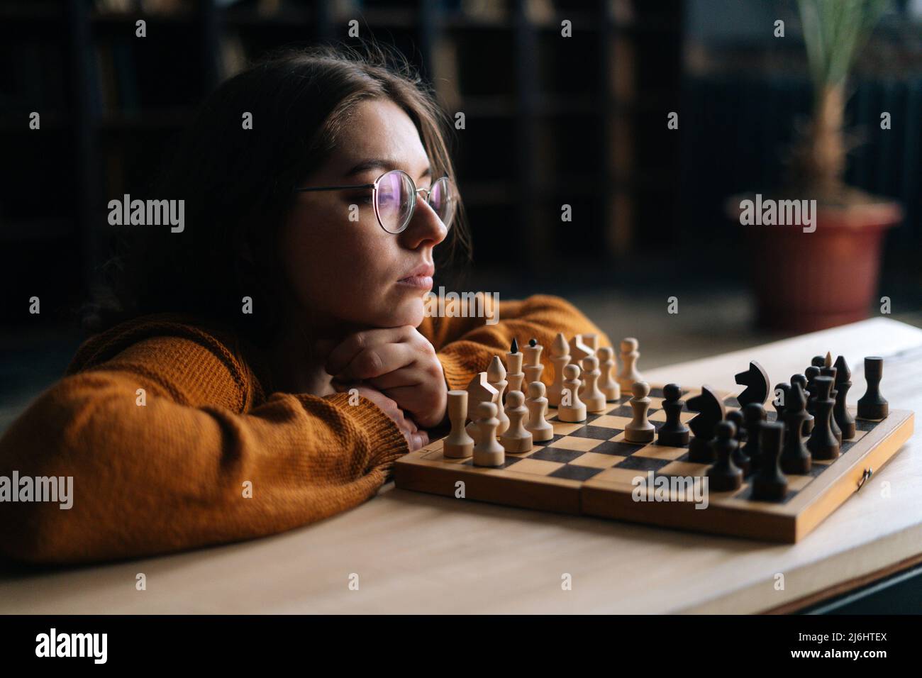 Nahaufnahme einer nachdenklichen Frau in eleganten Brillen, die an Schachbewegung denkt, die im dunklen Bibliotheksraum auf dem Boden sitzt, selektiver Fokus. Stockfoto