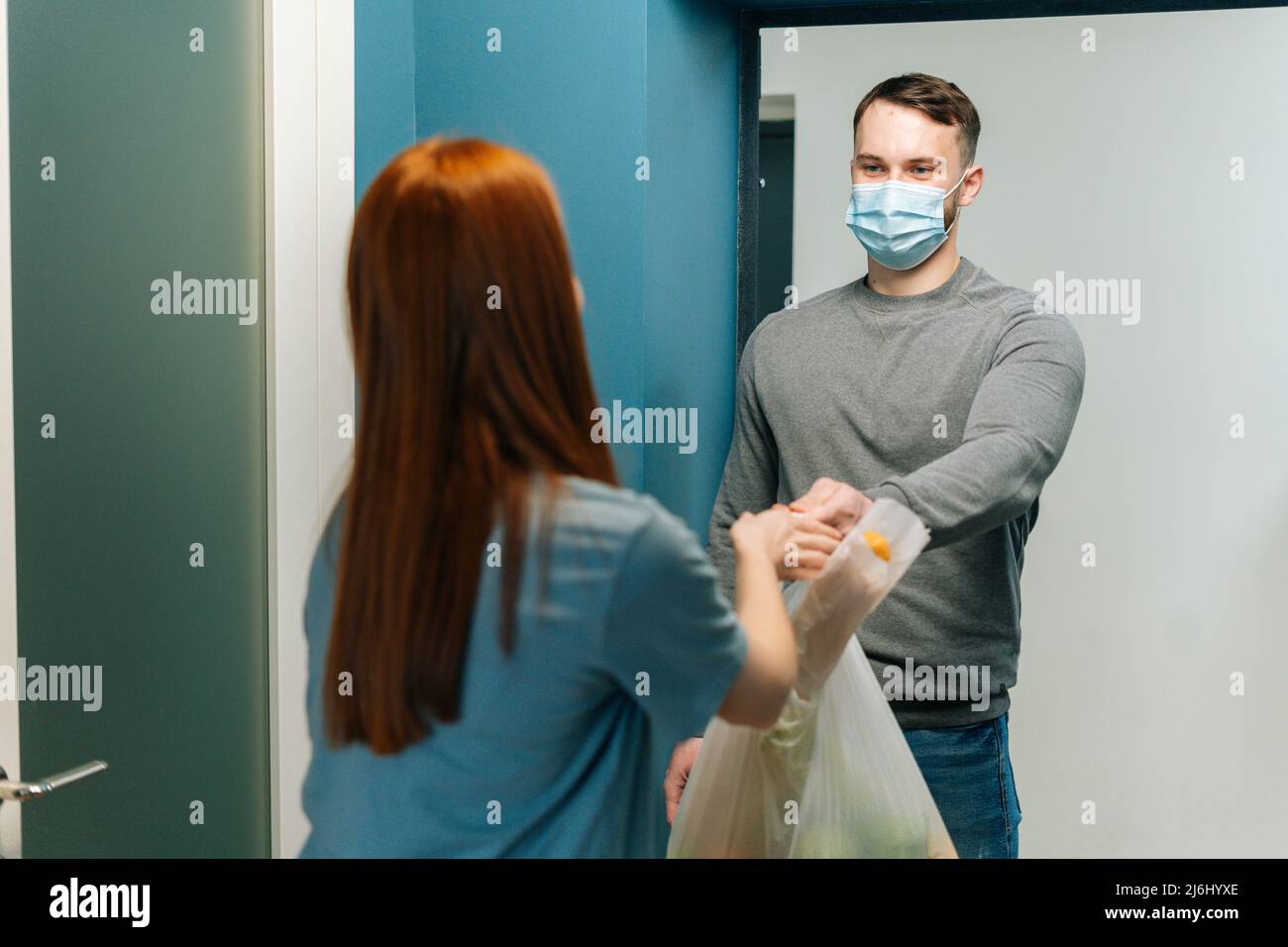 Hübscher Kurier männlich in schützender Gesichtsmaske liefert Lebensmittelbeutel an weibliche Hausfrau Kunde auf Tür zu Hause. Stockfoto