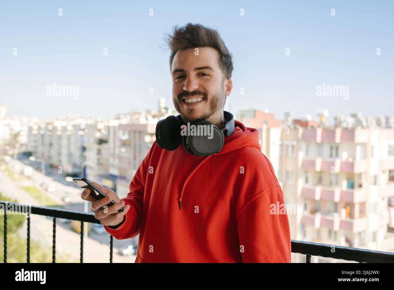 Posin, ein junger Gamer, der mit einem Telefon in der Hand für die Kamera posiert, nahm seine Kopfhörer ab und legte sie ihm auf den Hals, ein hübscher, erwachsener Mann, der lächelte Stockfoto