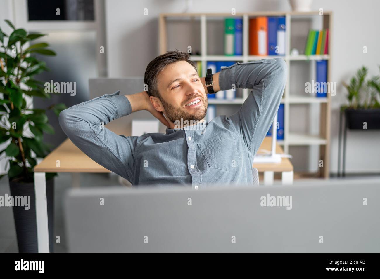 Erfolgreicher Geschäftsmann. Entspannter, reifer Mann, der die Hände hinter dem Kopf hält und am Schreibtisch im modernen Büro sitzt Stockfoto