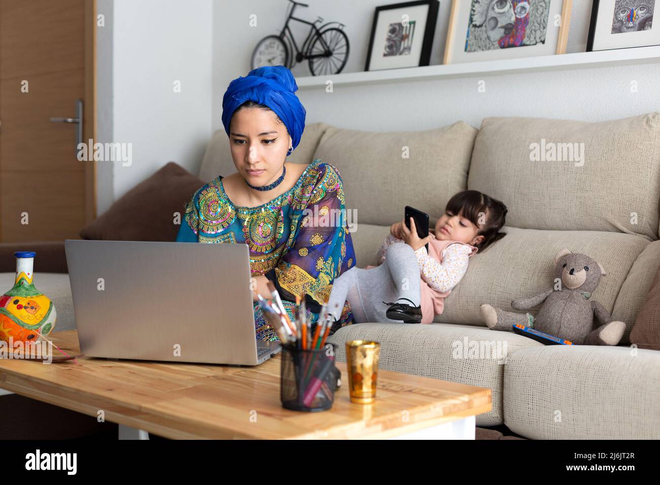 Junge muslimische Frau, die zu Hause mit ihrem Laptop arbeitet, während ihre junge Tochter mit einem Mobiltelefon spielt. Alleinerziehende Familie. Stockfoto