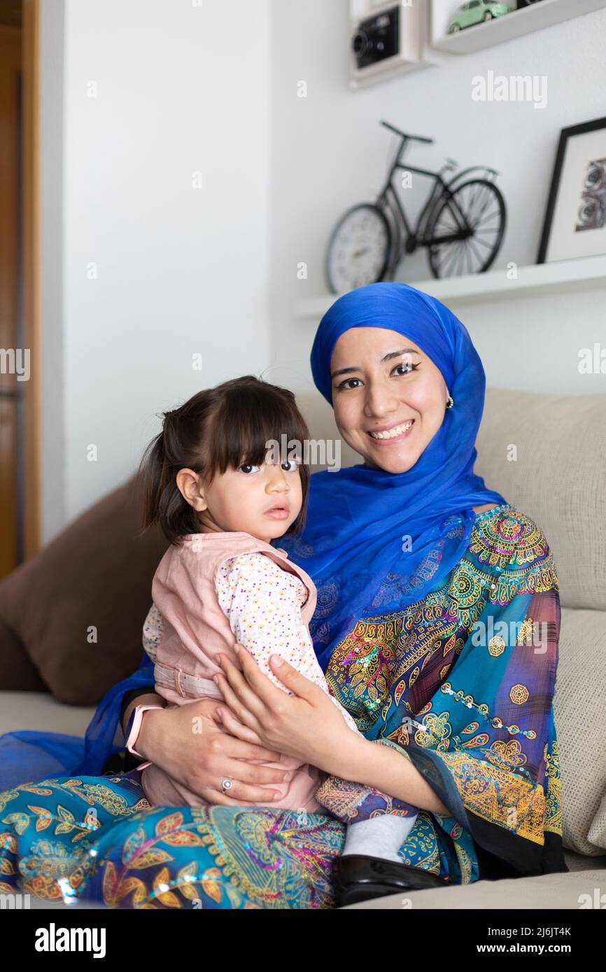 Junge muslimische Mutter sitzt auf dem Sofa mit ihrer kleinen Tochter in den Armen. Sie schauen auf die Kamera. Alleinerziehende Familie. Stockfoto
