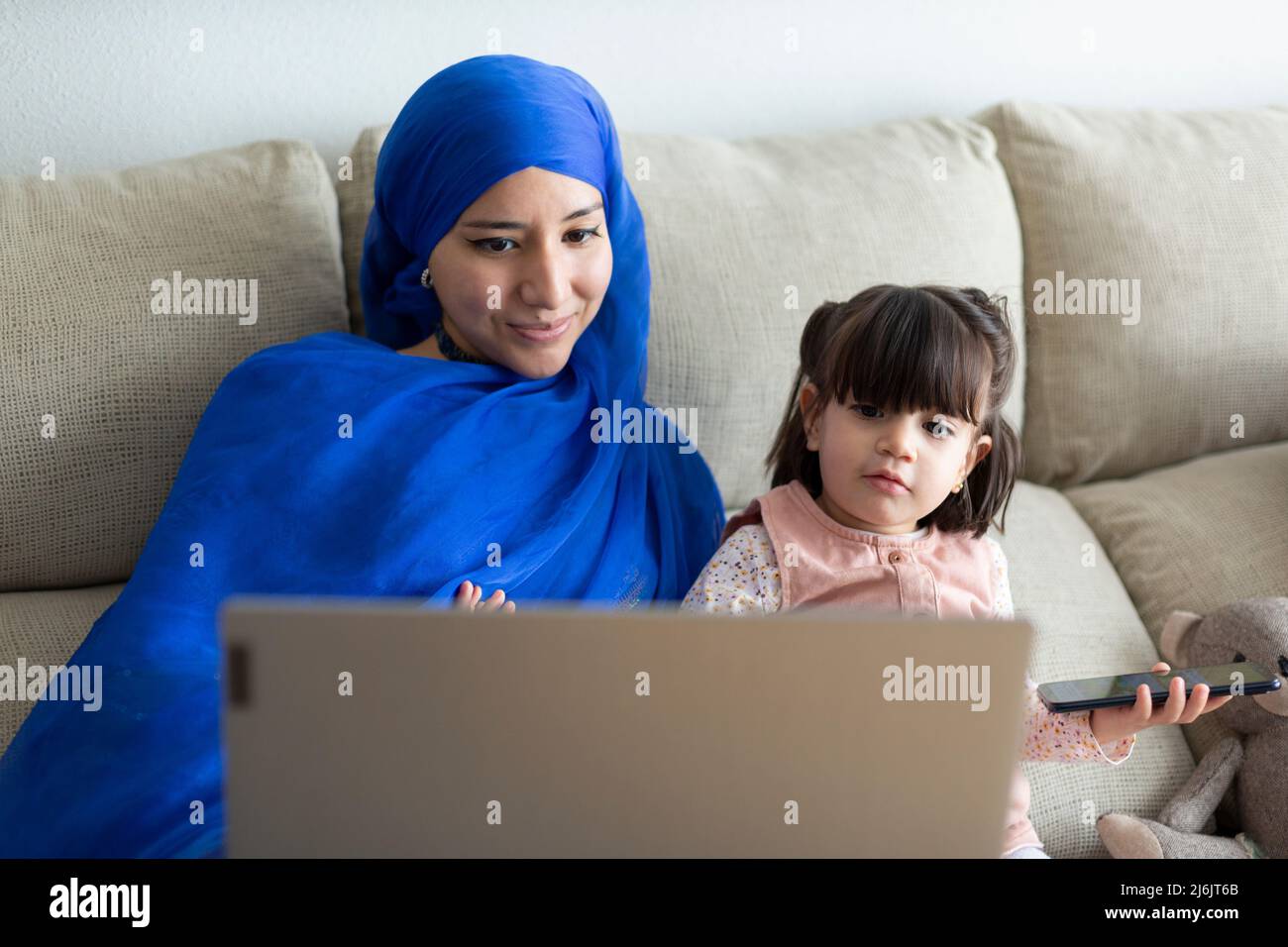 Junge alleinerziehende muslimische Mutter mit ihrer jungen Tochter, die zu Hause einen Laptop benutzt. Videoanruf tätigen, Online-Unterhaltung, Zeichentrickfilme zusammen ansehen und hav Stockfoto