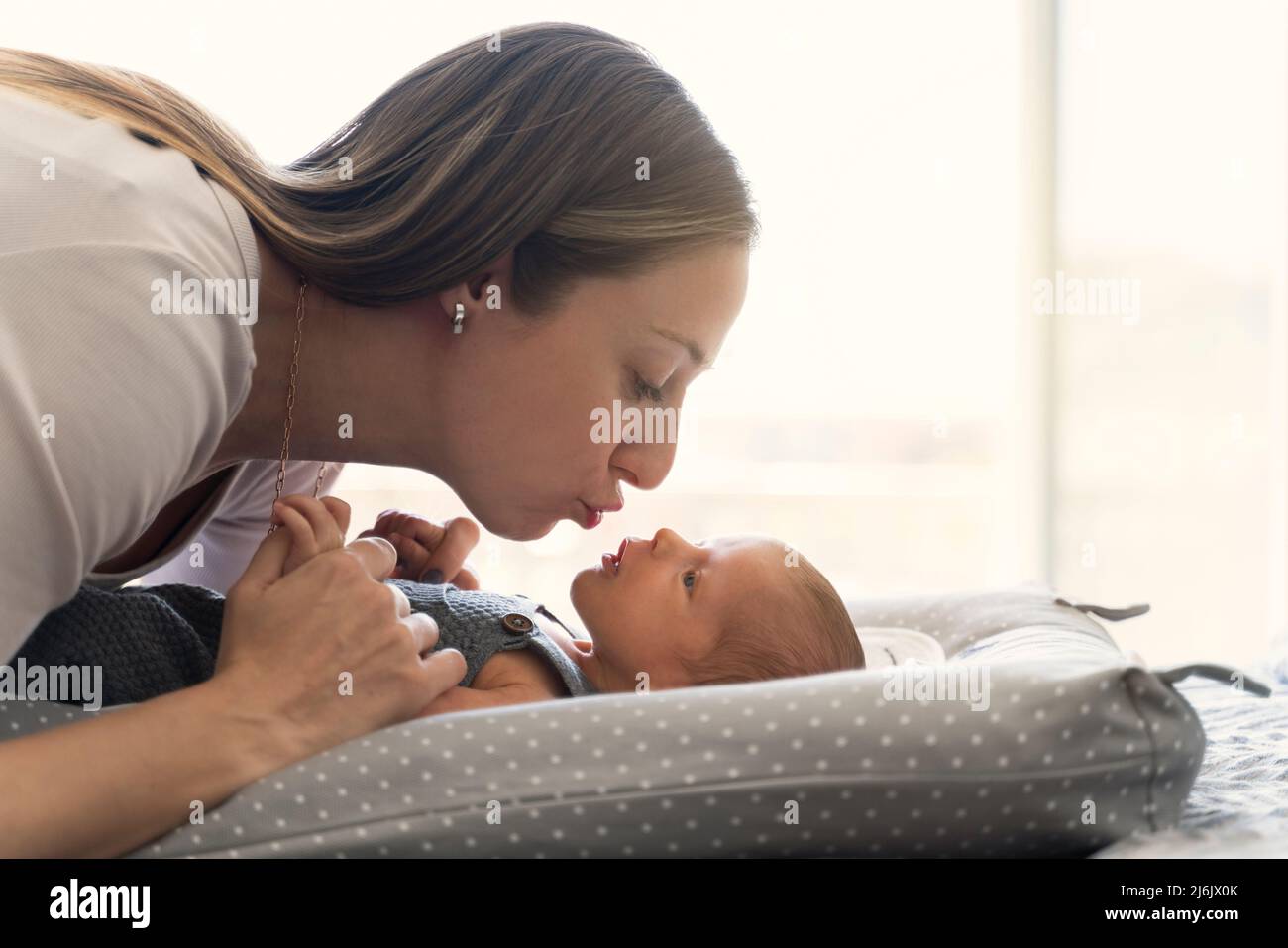 Die junge Mutter küsst ihr neugeborenes Baby auf dem Bett in der Nähe des Fensters Stockfoto