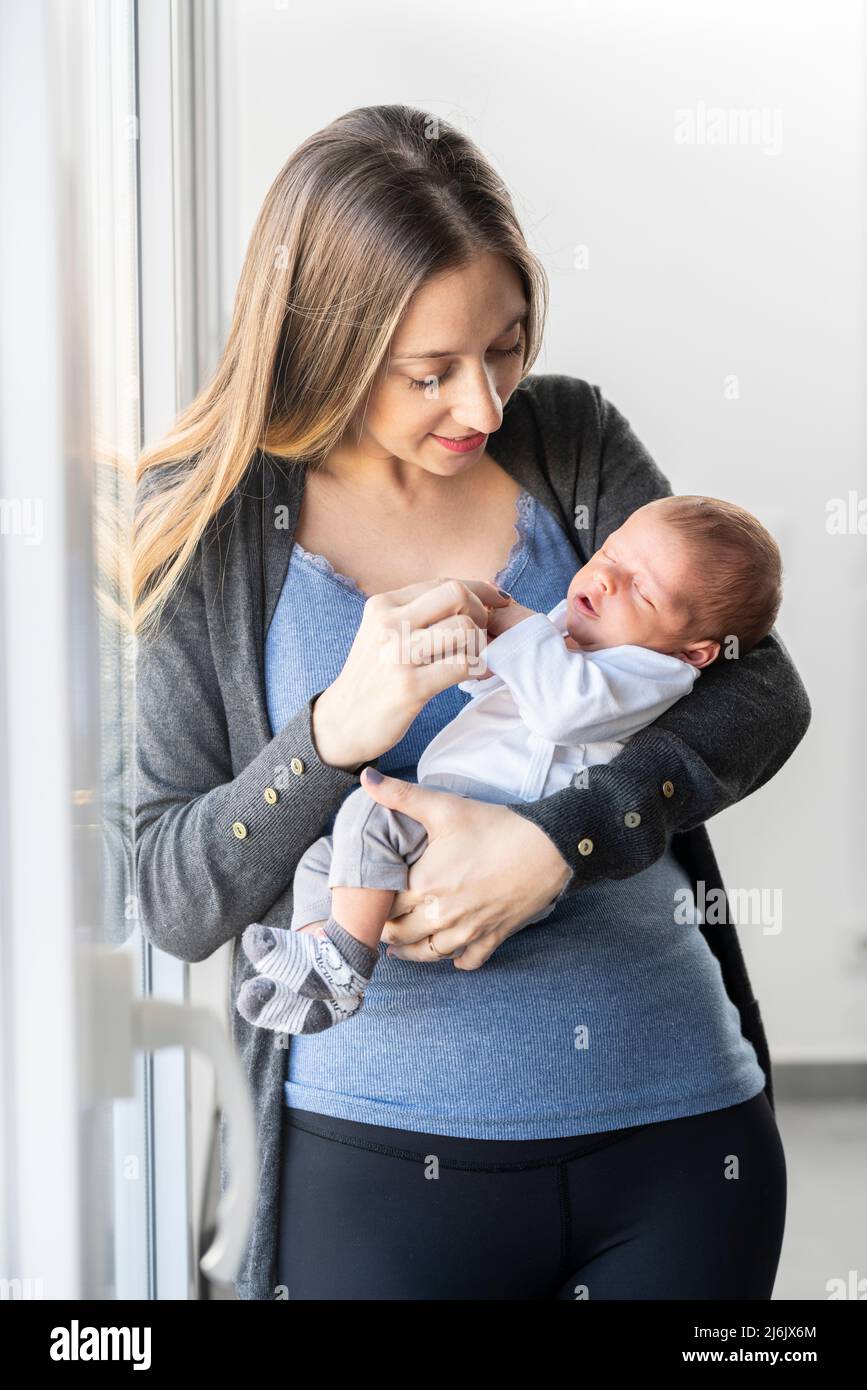 Junge Mutter mit ihrem neugeborenen Baby in den Armen zu Hause in der Nähe des Fensters Stockfoto