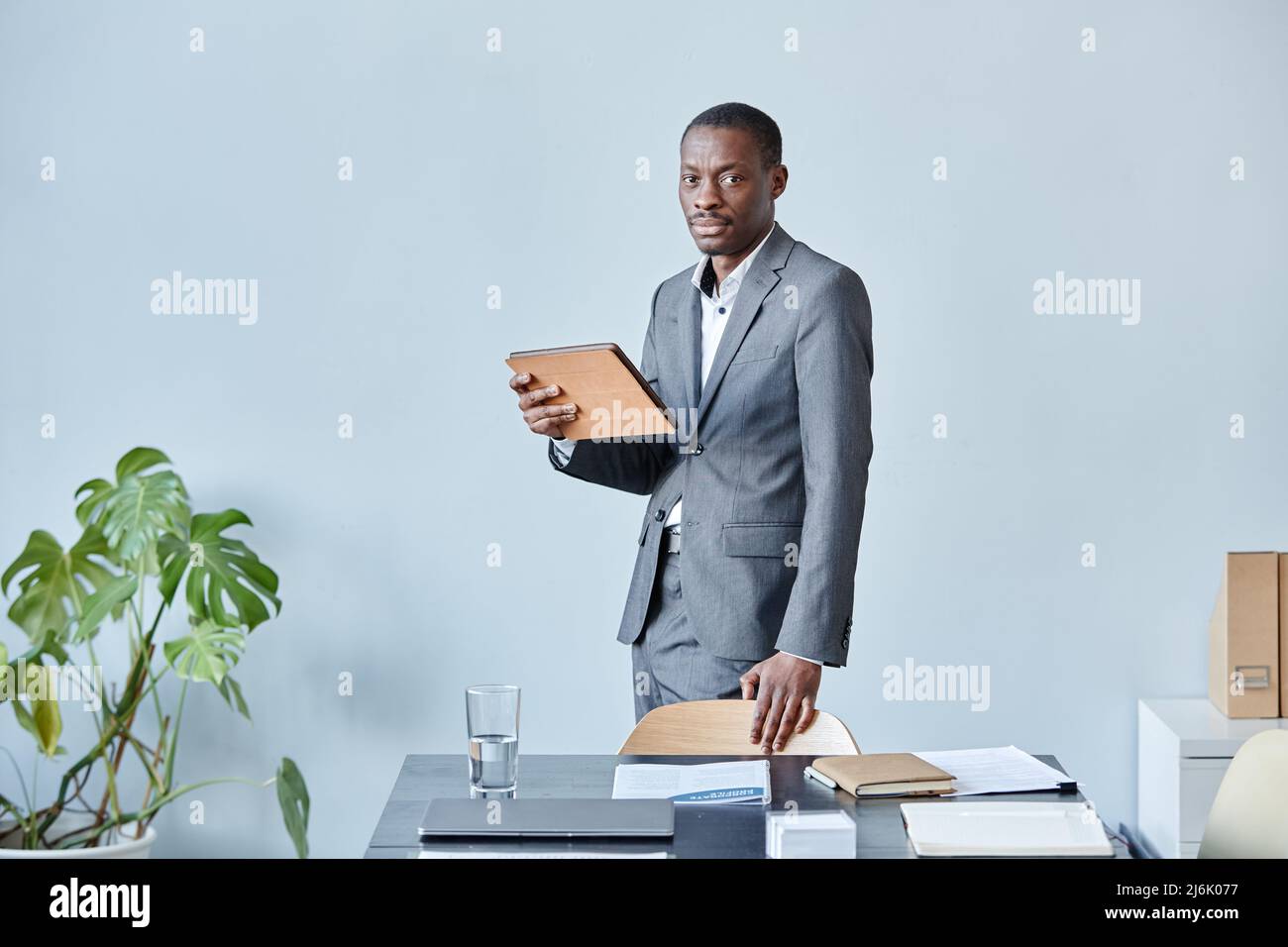 Minimales Porträt eines professionellen schwarzen Executive, der die Kamera anschaut, während er im Büro mit elegantem Anzug und Kopierraum gegen die blaue Wand steht Stockfoto