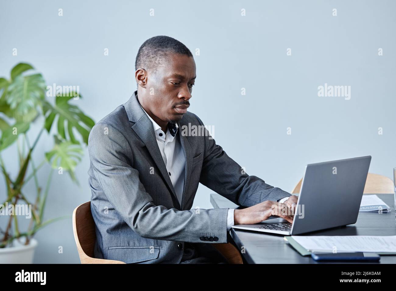 Minimales Porträt einer professionellen schwarzen Führungskraft, die einen Laptop verwendet, während sie am Arbeitsplatz im Büro gegen eine blaue Wand sitzt und einen Anzug trägt und Platz zum Kopieren verwendet Stockfoto