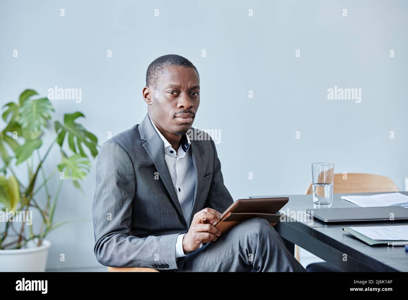 Minimalistisches Porträt eines professionellen schwarzen Executive, der die Kamera anschaut, während er am Arbeitsplatz im Büro gegen eine blaue Wand sitzt und einen eleganten Anzug, CO, trägt Stockfoto
