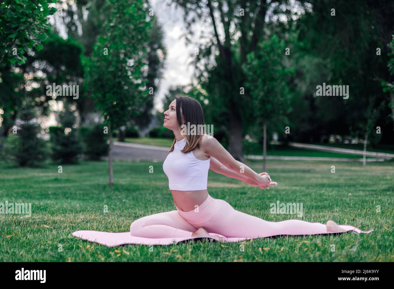 Junge athletische Frau praktiziert Yoga auf Gymnastikmatte im grünen Park auf Gras. Machen Sie Stretching-Übungen für eine gute Haltung in Sportbekleidung, freien Kopieplatz Stockfoto