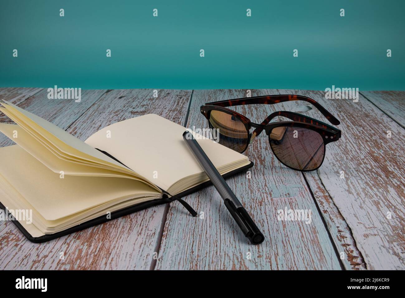 Notizbuch mit Stift und Sonnenbrille, auf einem Holztisch mit hellblauem Hintergrund. Konzept der freien Zeit. Stockfoto