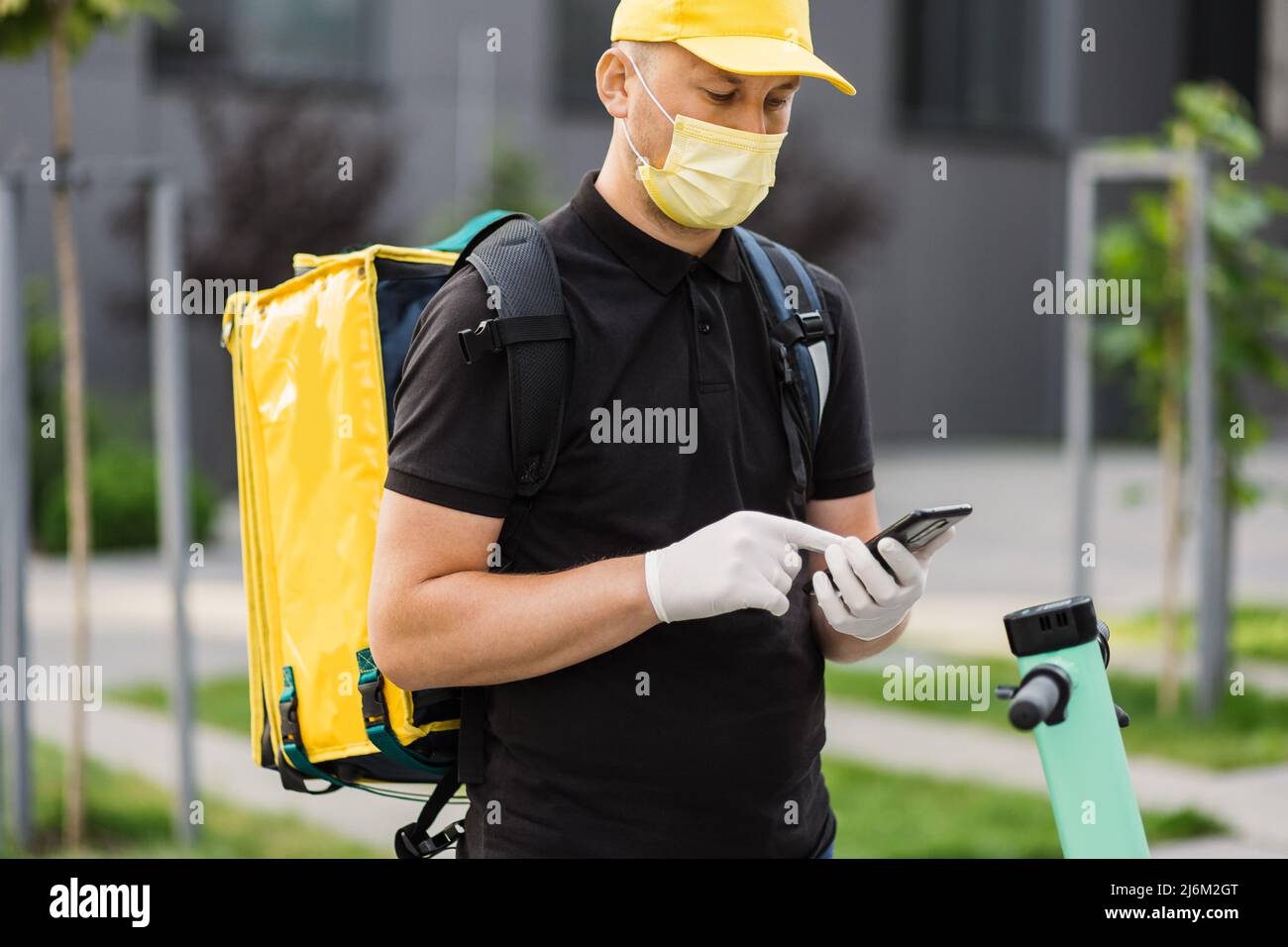 Nahaufnahme eines Kuriermanns in Gesichtsmaske, der im Mobiltelefon schaute, um die Adresse des Essens aus einem Restaurant zu liefern. Food Delivery Kerl navigiert im Telefon auf dem Hintergrund der Stadt mit Elektroroller. Stockfoto