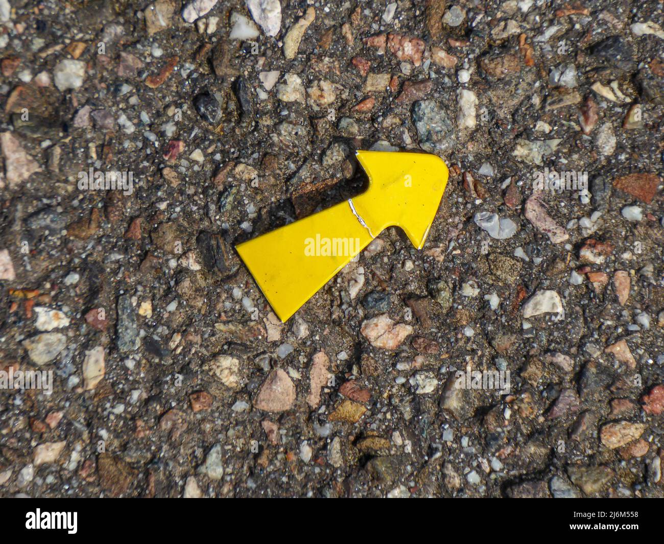 Ein kleiner, gelber Metallpfeil mit Gebrauchsspuren liegt auf einer rauen Fahrbahnoberfläche und zeigt nach rechts Stockfoto