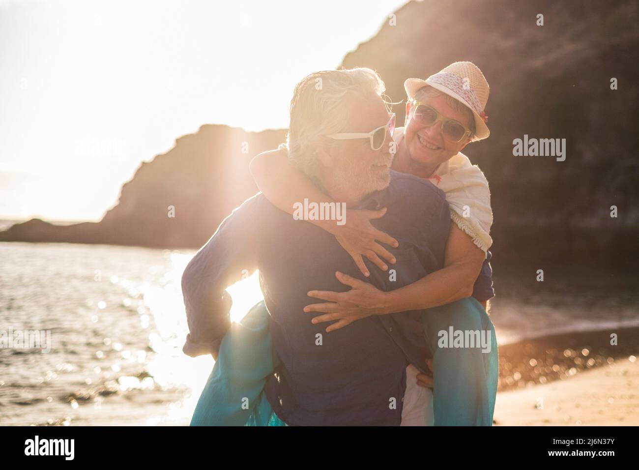 Alte Leute aktives Paar genießen Outdoor-Freizeitaktivitäten zusammen spielen und tragen Frau im Huckepack am Strand während Sonnenuntergang Urlaub Stockfoto