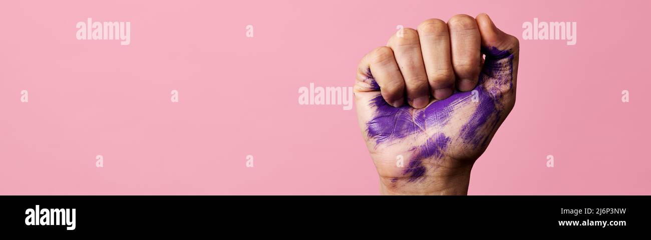 Nahaufnahme der erhobenen Faust eines Mannes mit einigen Flecken von violetter Farbe, auf einem rosa Hintergrund mit etwas leerem Raum auf der linken Seite, in einem Panoramaformat zu Stockfoto