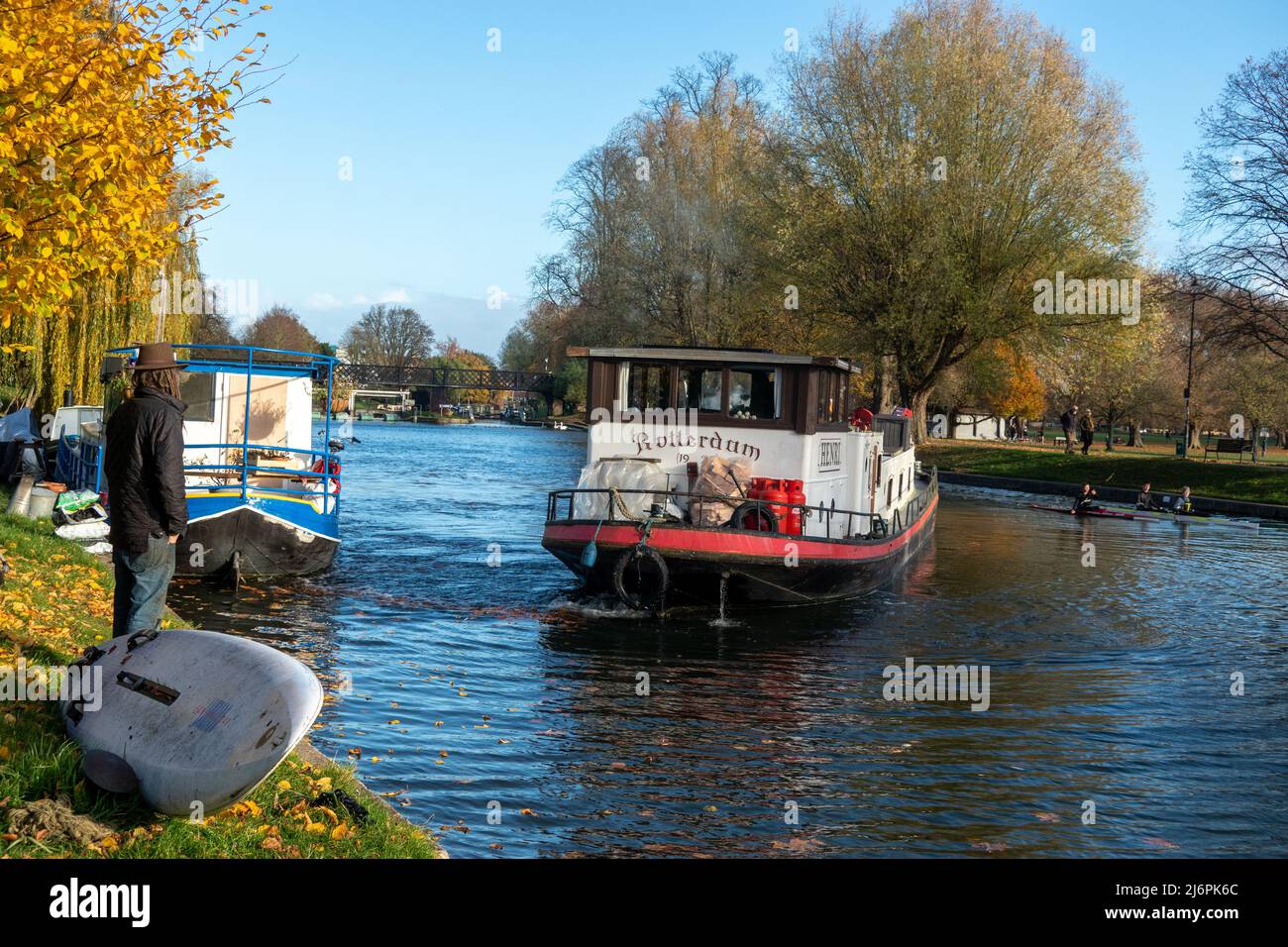Großes, schmal gefahrenen Boot und Kanus auf dem River Cam mit einem Mann, der am Flussufer neben dem Windsurfer in Cambridge, England, steht Stockfoto