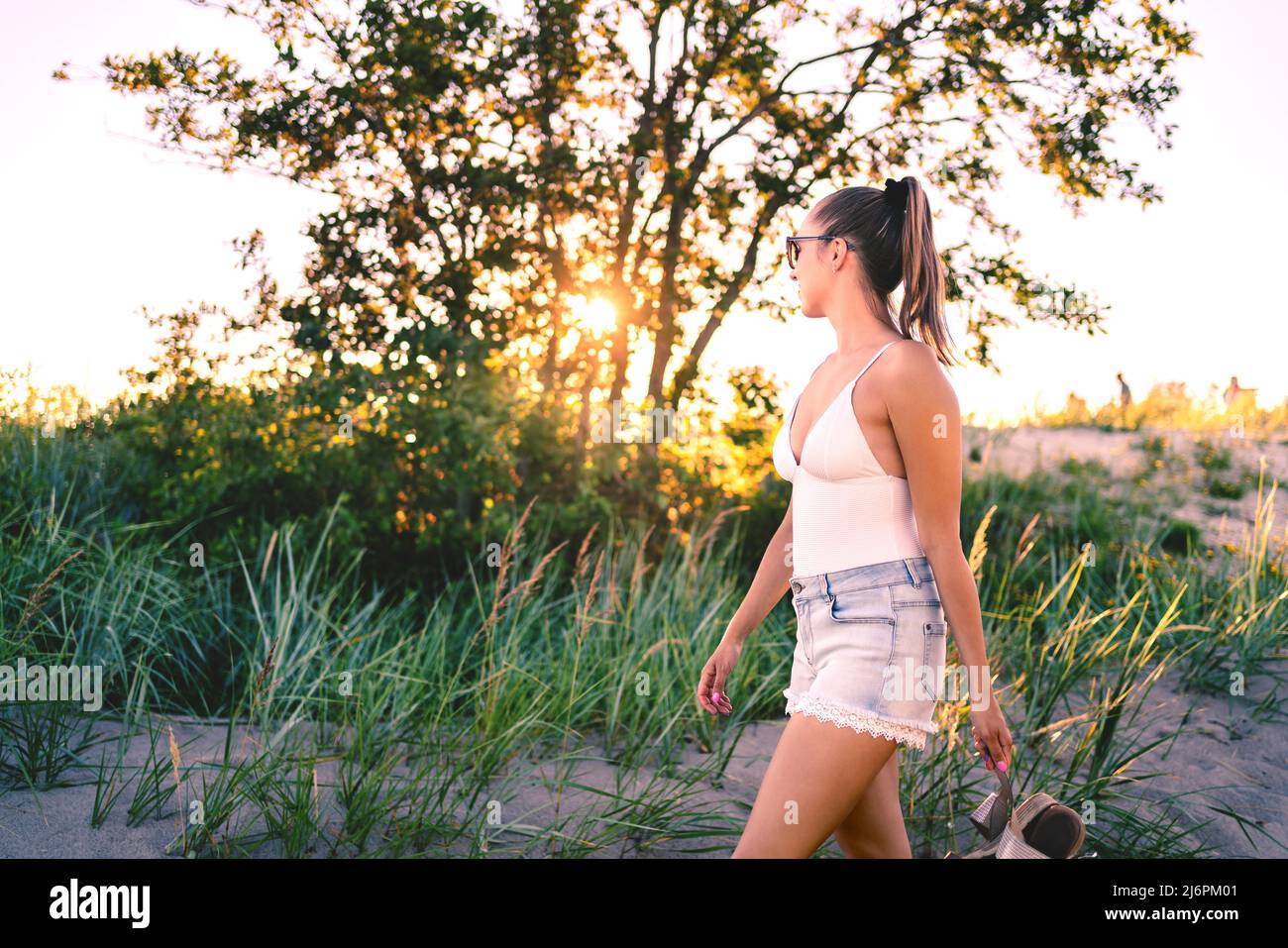 Sommerabend Spaziergang im Feld von Bäumen und Sanddünen bei Sonnenuntergang. Frau genießt friedliche Freizeit in der Natur im Freien. Trendiges, stilvolles Urlaubsoutfit. Stockfoto