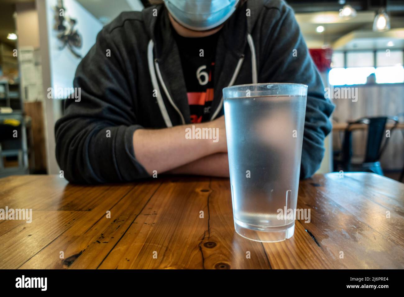 Blick aus der unteren Perspektive auf einen Mann in einer blauen Gesichtsmaske, der mit einem Glas Wasser an einem Holztisch in einem Restaurant sitzt Stockfoto