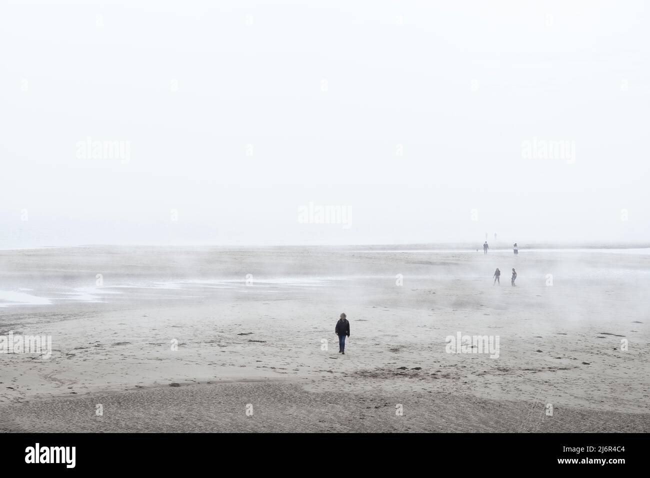 Eine Fotografin geht alleine auf die Kamera zu, mit einem flachen Sandstrand und einem Meeresnebel dahinter Stockfoto
