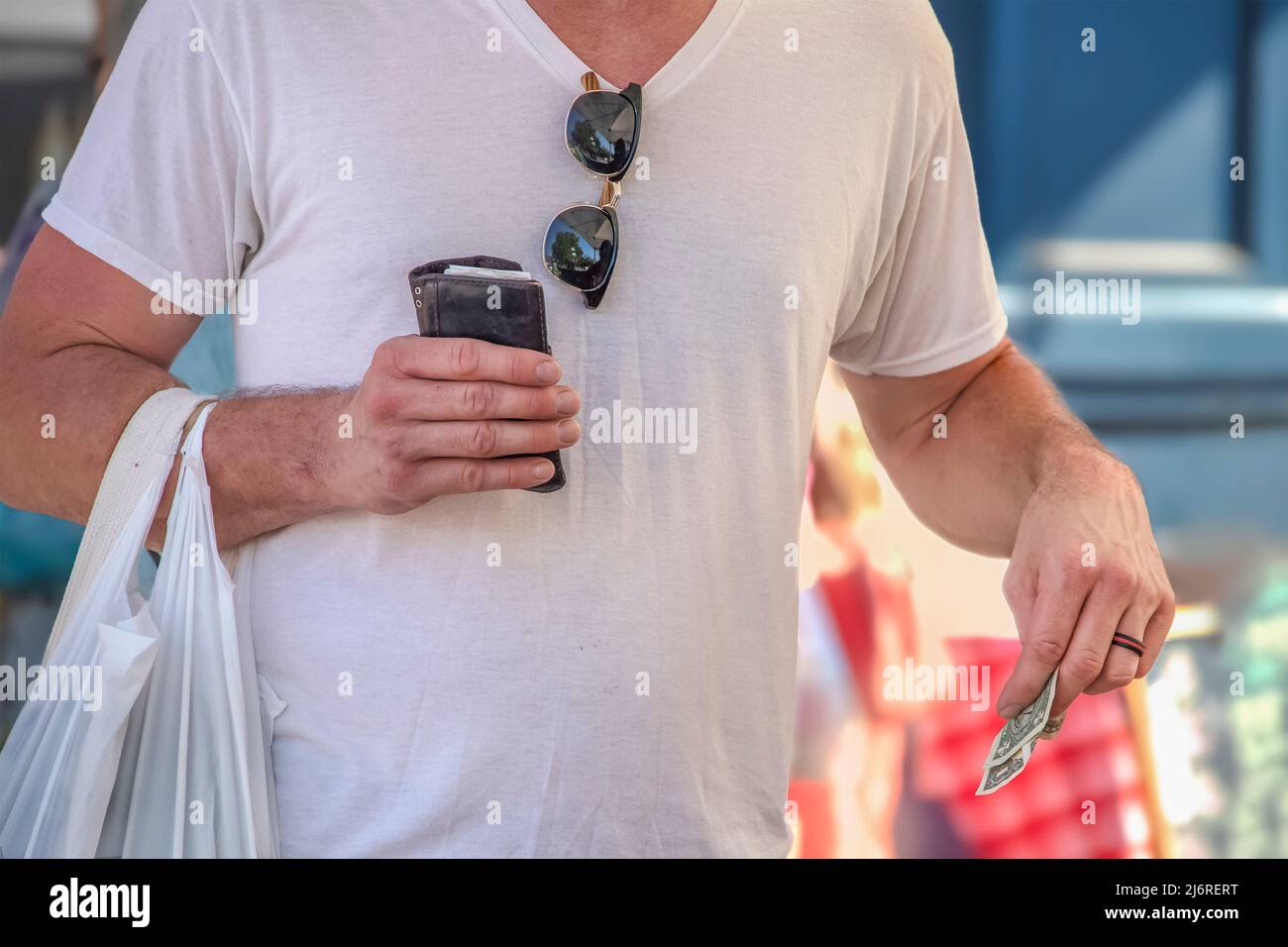 Ein Mann in weißem T-Shirt mit Sonnenbrille, die am Hals hing und eine Plastiktüte von dem hielt, was er gekauft und in Th mit Billfold und US-Dollar getragen hat Stockfoto