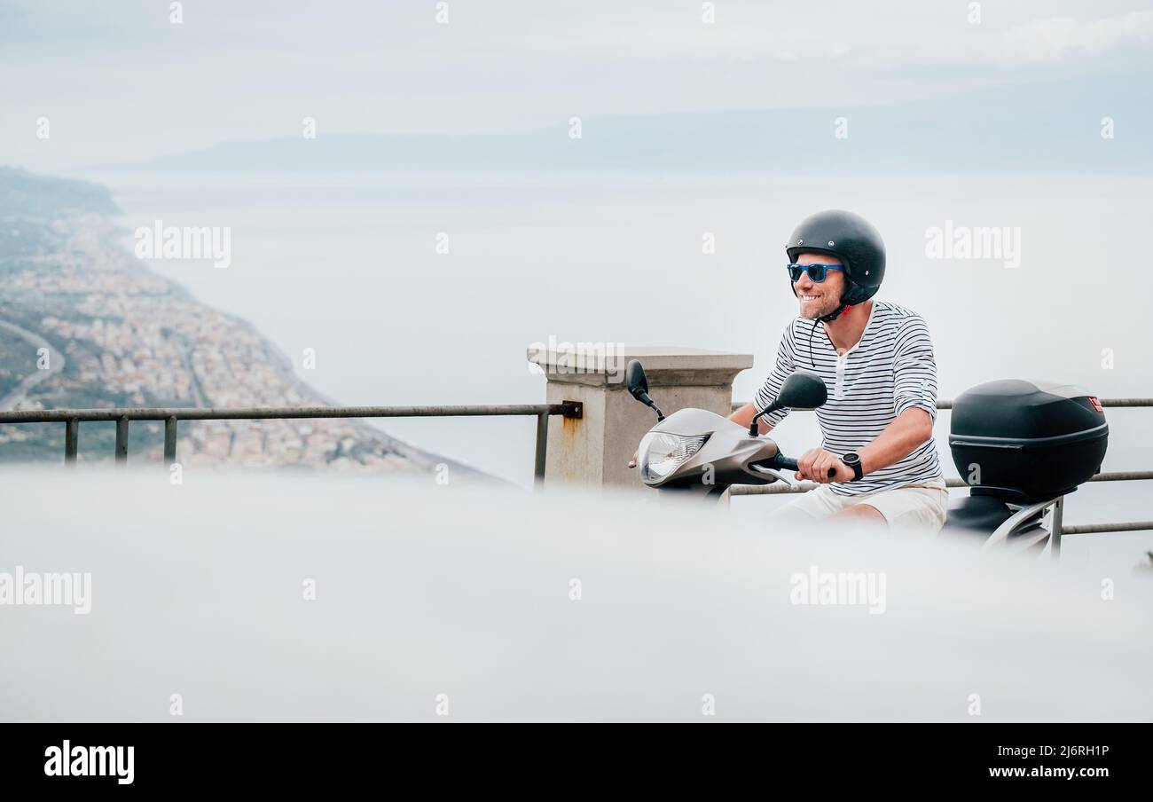Fröhlich lächelnder Mann mit Helm und Sonnenbrille fährt schnell mit dem Moto Scooter auf den Straßen der sizilianischen Altstadt an der ionischen Küste. Frohes Itali Stockfoto