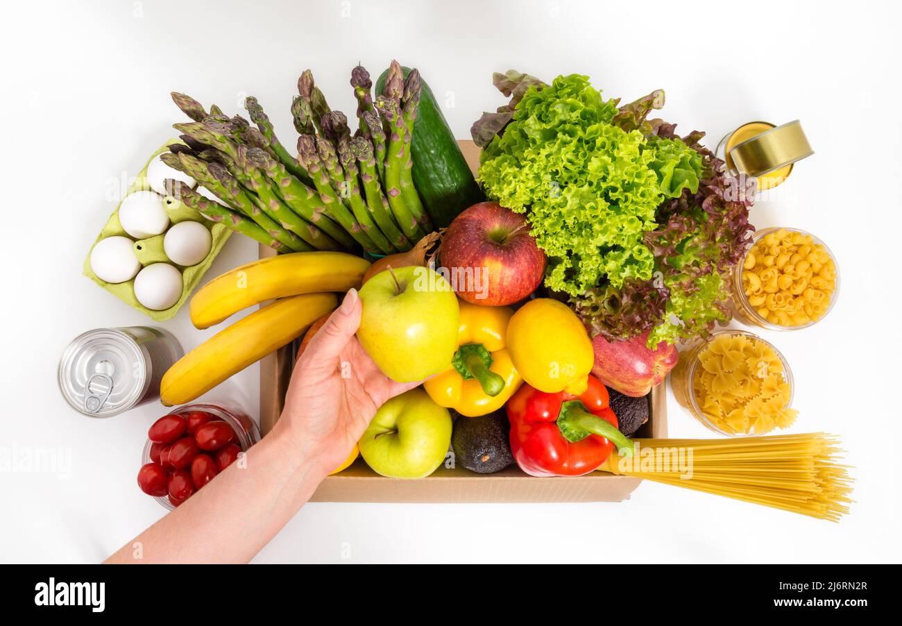 Weibliche Hand hält grünen Apfel über Box mit frischem Obst und Gemüse, Eier, Pasta und Konserven. Konzept der Spende oder Lieferung von Lebensmitteln. Vegetarisch Stockfoto