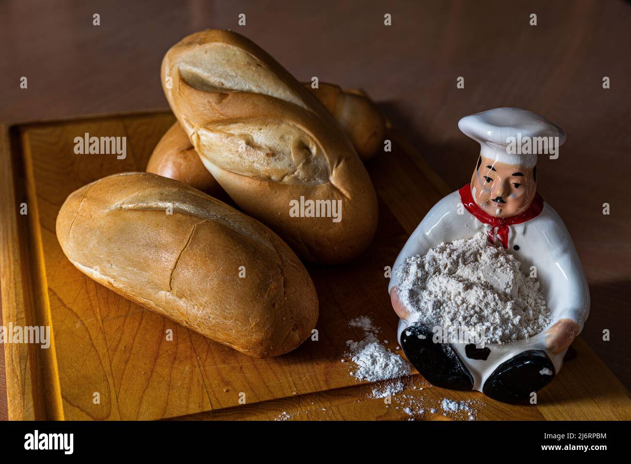 Frisches, leckeres Brot, Brötchen, ein Küchenmesser mit etwas Mehl auf einer Figur des Kochs, alles auf einem Schneidebrett. Stockfoto Frisches, leckeres Brot, Brötchen, ein Küchenmesser mit etwas Mehl auf einer Figur des Kochs, alles auf einem Schneidebrett. Stockfoto