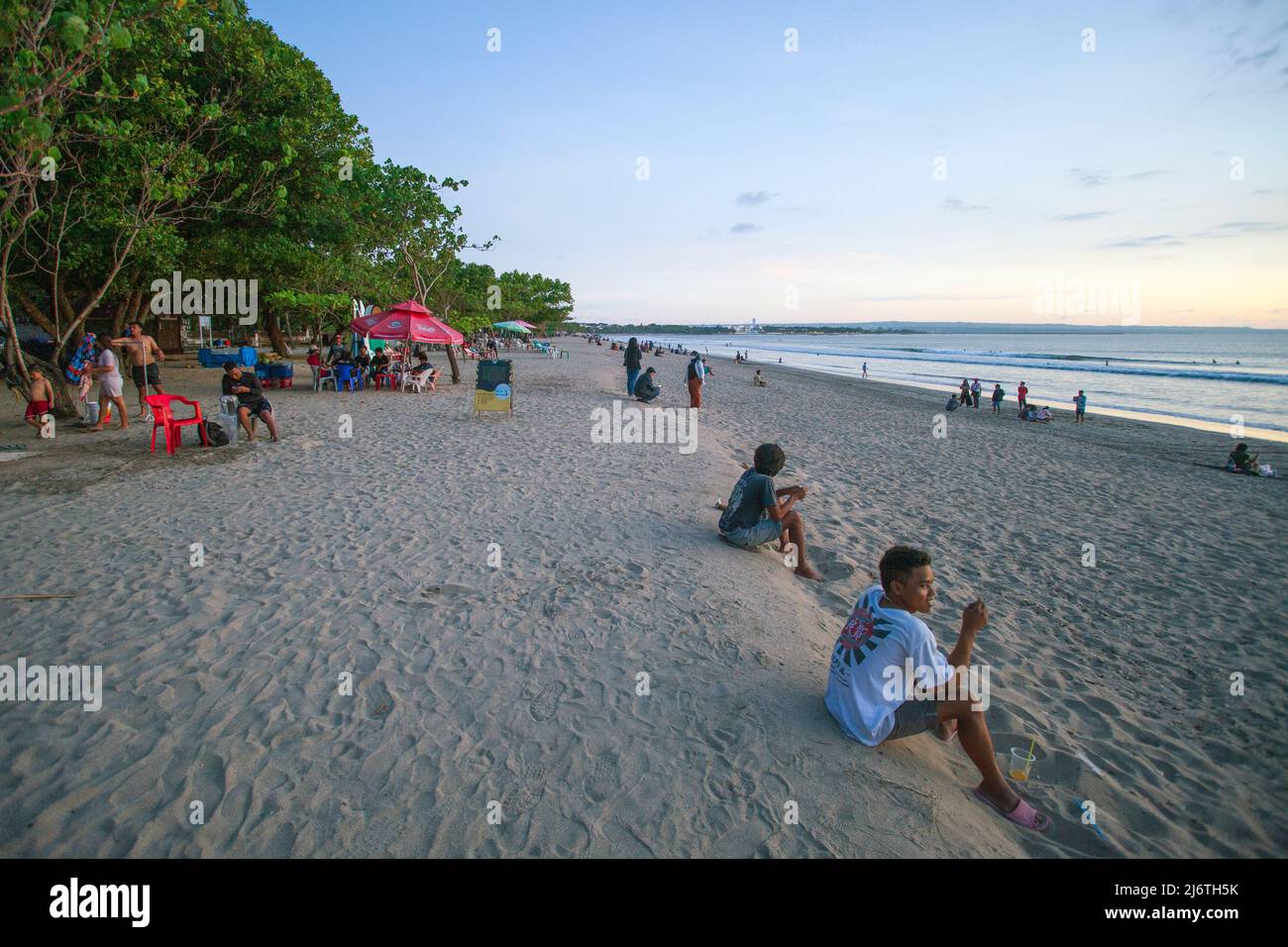 Blick auf den Strand von Kuta in Bali, wenn der Sonnenuntergang anrückt, während Menschen auf dem Sand und auf Stühlen auf den Sonnenuntergang warten. Stockfoto