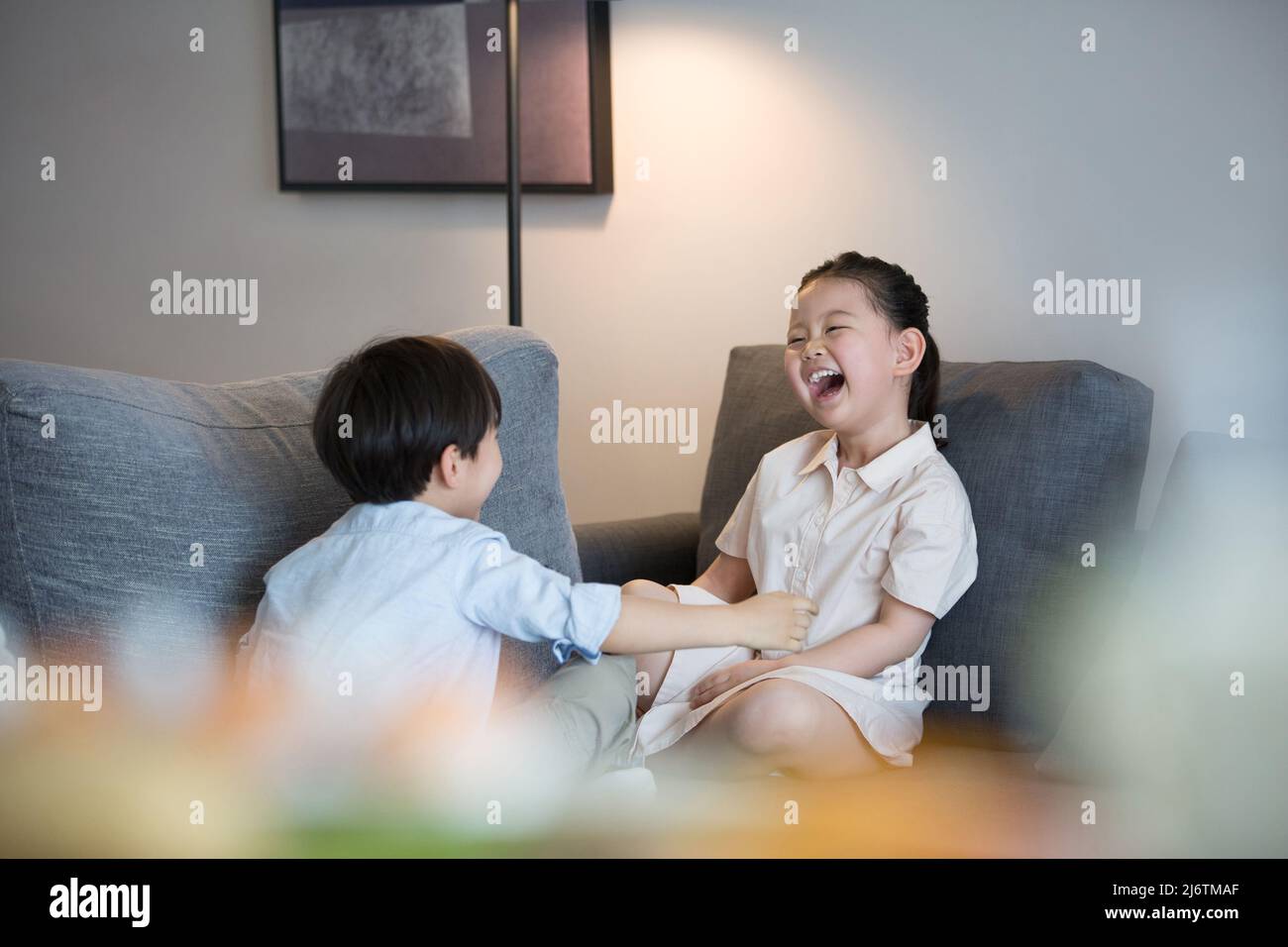 Ein kleines Mädchen und ein kleiner Junge sitzen mit gekreuzten Beinen auf dem Sofa im Wohnzimmer und lachen fröhlich - Stock photo Stockfoto